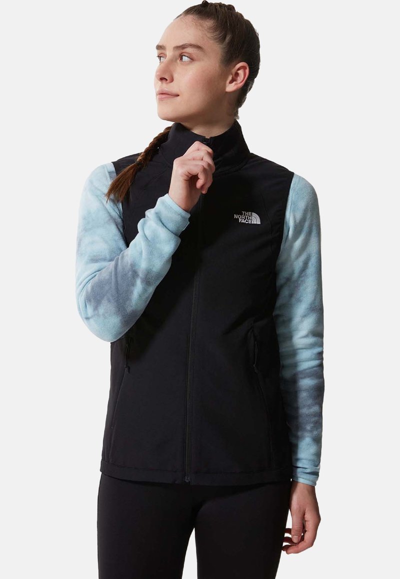Schwarze ärmellose Jacke mit zwei Seiten Taschen, getragen über einem hellblauen Batik Langarmshirt; verfügt über einen hohen Kragen und einen Frontreißverschluss.