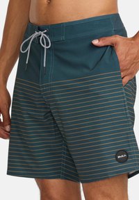 Männer Badehosen in Teal mit horizontalen goldenen Streifen. Ausgestattet mit einem Kordelzugverschluss und einem RVCA-Logo-Patch am Saum. Glattes, leichtes Material.