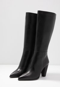 Botas de cuero negro hasta la rodilla con punta afilada y un tacón apilado de 7,5 cm. Textura suave con un detalle de costura mínimo a lo largo del eje.