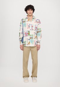 Valge nööbiga särk, millel on värvilised mustrid rannakaubast ja figuuridest. Kombineeritud beežide pükstega ja valgete tossudega.