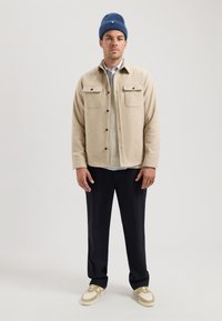 Beige uld skjortejakke med to brystlommer, lagret over en striktrøje. Marineblå beanie, sorte bukser og beige sneakers.