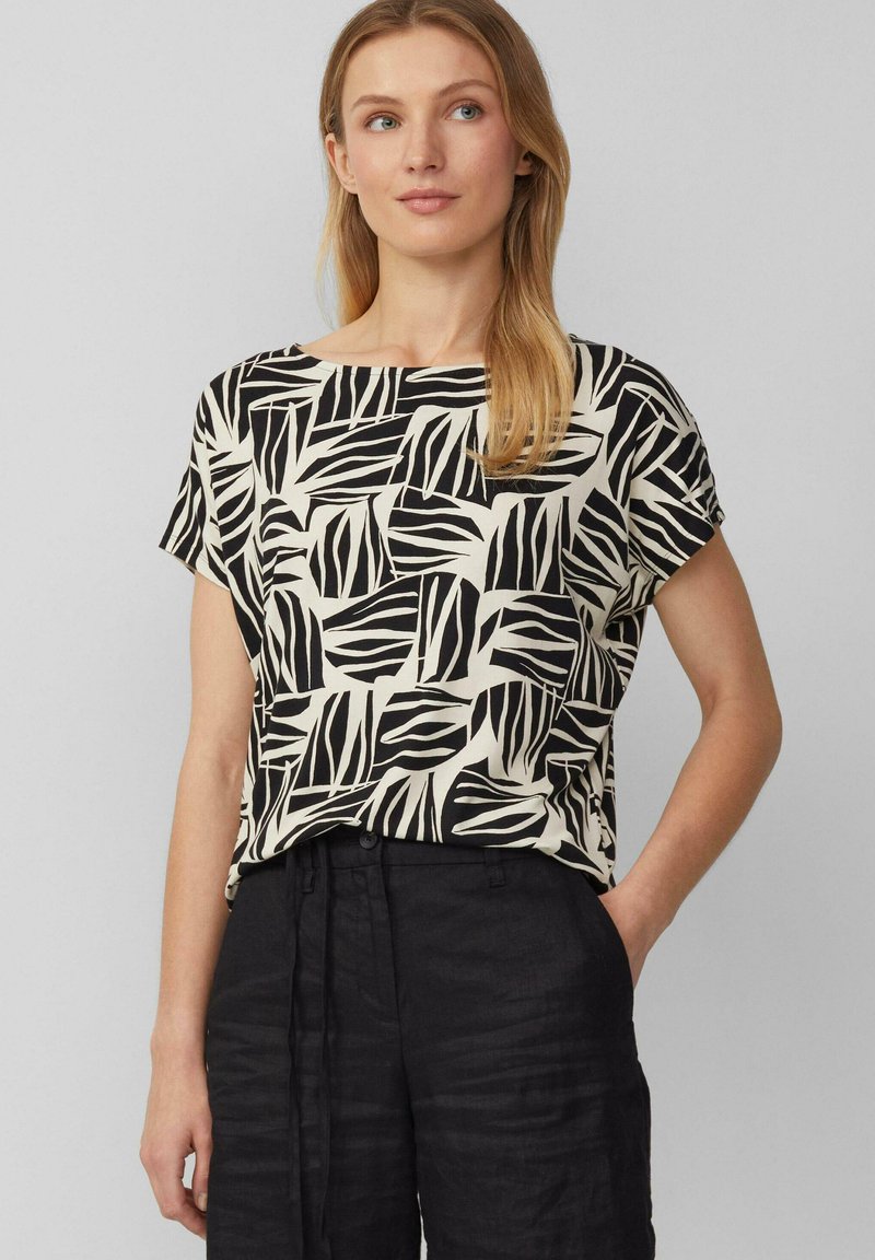 Korte mouwen blouse in crème met een gedurfde zwarte bladerenprint. Heeft een relaxte pasvorm en een ronde halslijn. Gecombineerd met zwarte linnen broek.