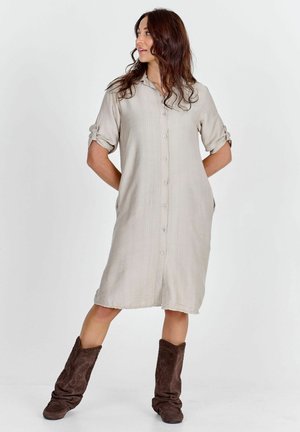 Femme debout, mains derrière le dos, portant une robe beige boutonnée avec manches retroussées et des bottes marron décontractées.
