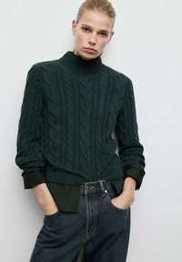 Suéter de tejido en patrón de cable de color verde oscuro con cuello alto y dobladillo acanalado, combinado con jeans de mezclilla oscuros de cintura alta. Patrón texturizado en todo el suéter.