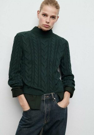 Suéter de tejido en patrón de cable de color verde oscuro con cuello alto y dobladillo acanalado, combinado con jeans de mezclilla oscuros de cintura alta. Patrón texturizado en todo el suéter.
