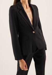 Black blazer with a deep V-neck, structured shoulders, single gold floral button, two front pockets, and smooth fabric texture.