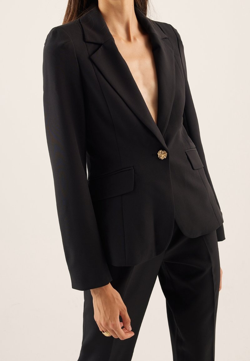 Black blazer with a deep V-neck, structured shoulders, single gold floral button, two front pockets, and smooth fabric texture.