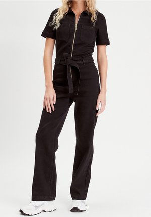 Kobieta ubrana w czarny kombinezon z krótkim rękawem, z suwakiem z przodu, paskiem wiązanym w talii i białe trampki, stojąca na białym tle.