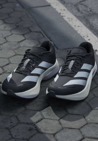 Paire de chaussures de course Adidas Adizero noir et blanc posées sur un pavé hexagonal près d'un trottoir.