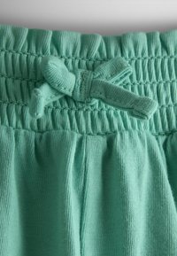 Primo piano di pantaloni lavorati a maglia color verde menta con vita elasticizzata arricciata e fiocco di cordoncino annodato al centro frontale.