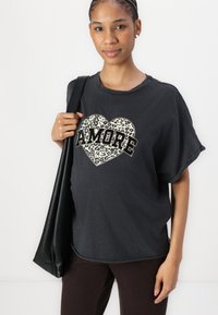 Černé oversized tričko s leopardím potiskem ve tvaru srdce a nápisem "AMORE" tučným písmem. Kombinováno s tmavě hnědými kalhotami a černou kabelkou.