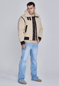 Beige shearling jacket with brown leather accents and black ribbed cuffs; features a large collar and zipper closure, paired with light blue jeans.