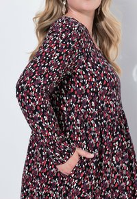 Schwarzes Langarmkleid mit einem bunten Muster aus roten, weißen und lavendelfarbenen Formen; verfügt über einen gerafften Rock und seitliche Taschen.