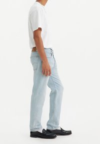 Jeans de denim azul claro con corte recto, que presentan una etiqueta de cuero en la parte posterior, combinados con una camiseta blanca y mocasines negros.