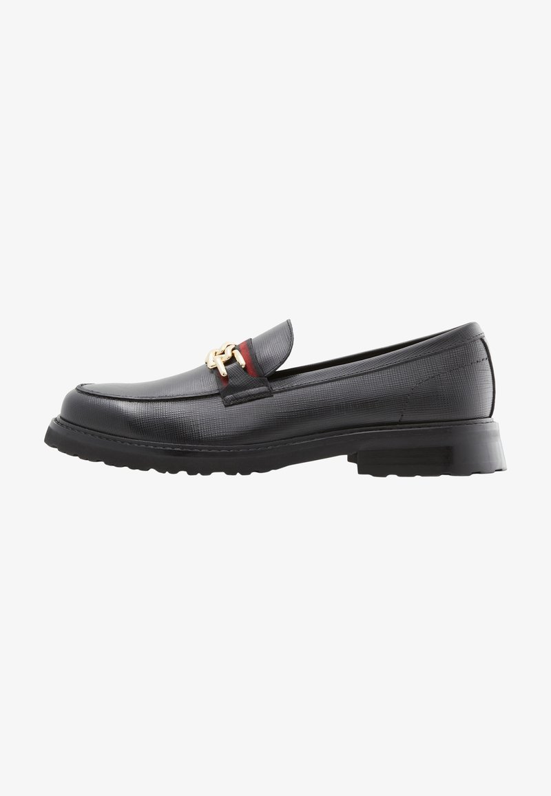 Mocassin en cuir noir avec un détail chaîne dorée et une discrète bande rouge sur le dessus, doté d'une semelle épaisse crantée.