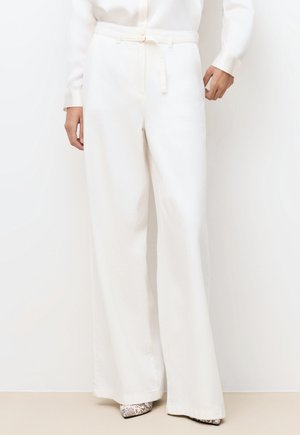 Frau trägt hoch taillierte, weit geschnittene weiße Hose mit passendem dünnen Gürtel und spitz zulaufenden Schuhen im Schlangenledermuster.