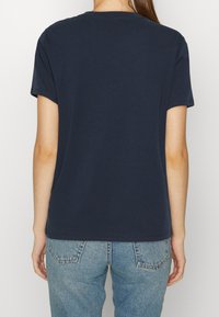 Kvinna som bär en enkel mörkblå kortärmad t-shirt och ljusblå denimjeans, sedd bakifrån mot en vit bakgrund.