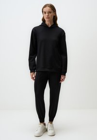 Czarna, oversize bluza z kapturem i sznurkiem, połączona z czarnymi joggerami o zwężanym kroju z elastycznymi mankietami. Model ma na sobie białe sneakersy.