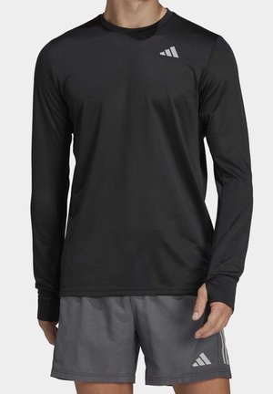 Mann trägt schwarzes langärmeliges Sportshirt und graue Adidas-Shorts mit weißem Logo, steht vor einem einfachen Hintergrund.