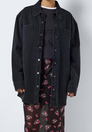 Person iført en sort oversized skjorte med knapper over en sort top og et gennemsigtigt sort skørt med røde og hvide blomster.