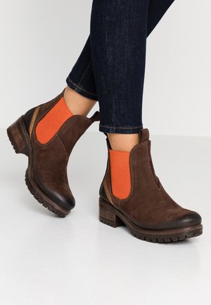 Bottes cheville en cuir marron avec panneaux latéraux élastiques orange portées avec un jean bleu foncé sur un fond uni.