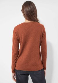 Frau mit langen braunen Haaren, die einen rostfarbenen Longsleeve-Pullover und dunkle Hosen trägt, mit dem Rücken zu einem schlichten weißen Hintergrund.