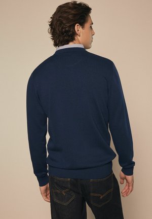 Marineblaues Strickpullover mit Rundhalsausschnitt und gerippten Bündchen, getragen über einem Hemd mit Kragen, kombiniert mit dunkelblauen Jeans.