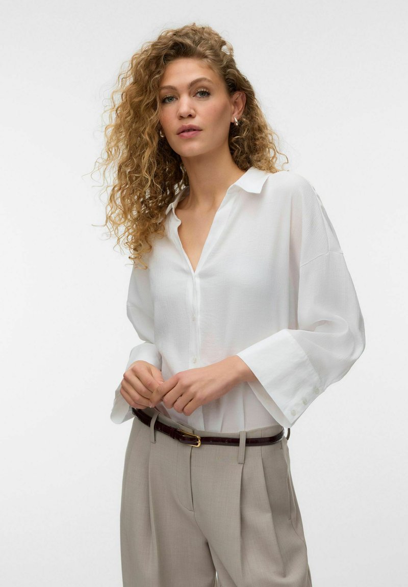 Jeune femme aux cheveux bouclés portant une chemise blanche ample boutonnée et un pantalon beige, ajustant sa ceinture foncée sur un fond uni.