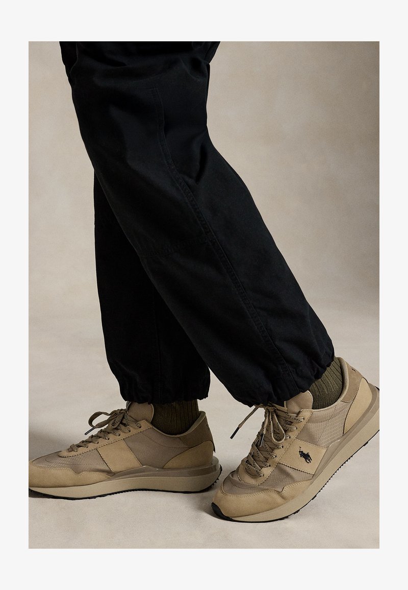 Beige sneakers with black logo worn with olive socks and black elastic-cuffed pants against a neutral background.