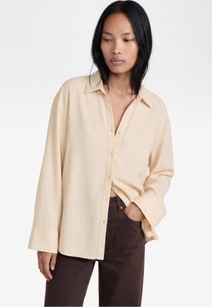 Kvinde med langt sort hår iført en oversized beige skjorte med knapper og mørkebrune bukser, stående med den ene hånd i lommen og kiggende nedad.