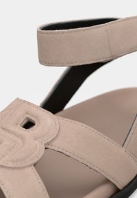 Μπεζ σουέτ σανδάλι με δύο σταυρωτούς ιμάντες, ρυθμιζόμενο λουράκι αστραγάλου, στρογγυλή τρύπα στον ιμάντα και αναπαυτική εσωτερική σόλα με μαύρη σόλα.