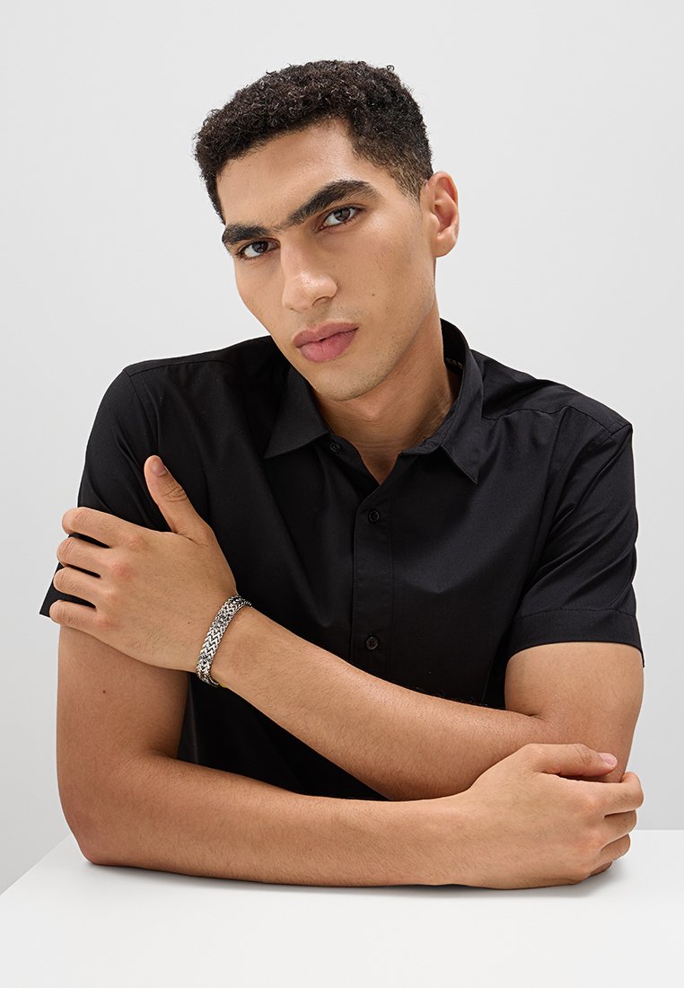 Černá košile s krátkým rukávem, hladká textura, viditelné knoflíky. Stříbrný pletený náramek na zápěstí, štíhlé prsty překřížené přes ruku.