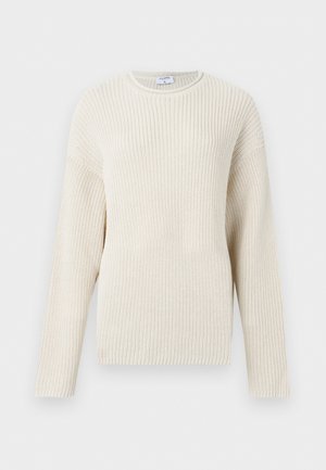 Kvinde står med hånden i lommen, iført cremefarvet striktrøje, lysegrønne shorts og beige spidse flade sko mod en ensfarvet hvid baggrund.