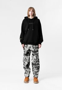 Sudadera negra con capucha y detalle en el panel frontal, combinada con pantalones de patrón en blanco y negro. Zapatos marrones con suelas texturizadas.