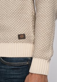 Knit sweater featuring a beige textured pattern with a light beige ribbed hem. Includes a brown leather logo patch on the lower side.