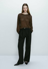 Jeune femme portant un pull en laine marron, un pantalon large noir avec une ceinture tressée, et des chaussures noires à bout pointu, se tenant devant un fond uni.