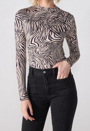 Mujer con una blusa de manga larga beige con rayas negras de cebra, metida dentro de unos vaqueros negros de talle alto, con una mano en el bolsillo.