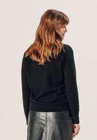 Zwarte gebreide trui met een geribde onderrand, ronde hals en slanke manchetten, gedragen met een zwarte leren rok, gezien vanuit de achterkant.
