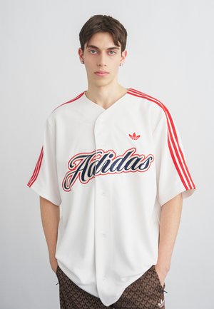 Νέος άνδρας που φοράει μια λευκή φανέλα Adidas με κόκκινες ρίγες και λογότυπο σε γραφή, στέκεται με τα χέρια στις τσέπες καφέ παντελονιού με σχέδια.