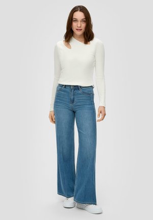 Haut blanc à manches longues avec un design découpé au niveau de l'épaule, associé à un jean taille haute à jambes larges bleu et des baskets blanches.