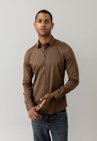 Camisa marrón de manga larga con cuello puntiagudo, botones al frente y textura suave, combinada con jeans azules. El modelo gesticula con las manos.