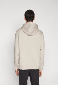 Beige hoodie med dragsko i huven och ribbade muddar, tillverkad av mjukt tyg, ihop med mörka cargobyxor. Bakifrån finns det inga grafiska detaljer.