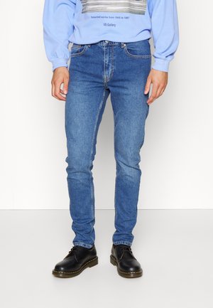 Personne portant un jean bleu slim, des chaussures en cuir noires à lacets et un sweat-shirt bleu clair avec texte et image sur le devant.