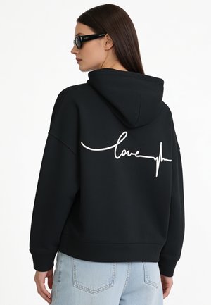 Vrouw met lang donker haar, die een zwarte hoodie draagt met witte "love" en een hartslijnontwerp op de achterkant, lichtblauwe spijkerbroek en zwarte zonnebril.