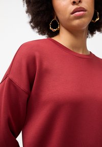 Felpa color marrone scuro con scollatura a girocollo e vestibilità ampia. Presenta un tessuto liscio e texturizzato e un design con spalle scese. Orecchini a cerchio dorati visibili.