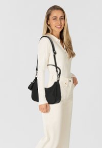 Zwarte leren crossbody tas met een gebogen vorm en een gestructureerd oppervlak. Verstelbare band met zilveren hardware. Draagt door een model in lichte kleding.