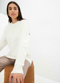 Pullover bianco soffice con colletto rotondo e polsini a coste, abbinato a pantaloni beige lucenti, seduta su uno sgabello di legno contro uno sfondo chiaro.