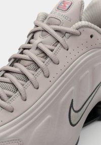 Nike Shox ketsid heledas roosas, tekstureeritud nahast ülemise osaga, silmapaistvate hallide paeltega ja hõbedase Nike swoosh logo külje peal.