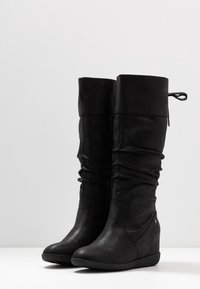 Bottes noires montantes en matériau synthétique lisse, présentant un design décontracté, une fermeture éclair et des talons compensés avec semelles texturées.