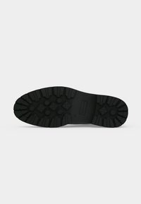 Semelle de botte en caoutchouc noir avec des bandes de roulement profondes et des motifs d'adhérence circulaires et en forme de croix sur un fond gris clair uni.